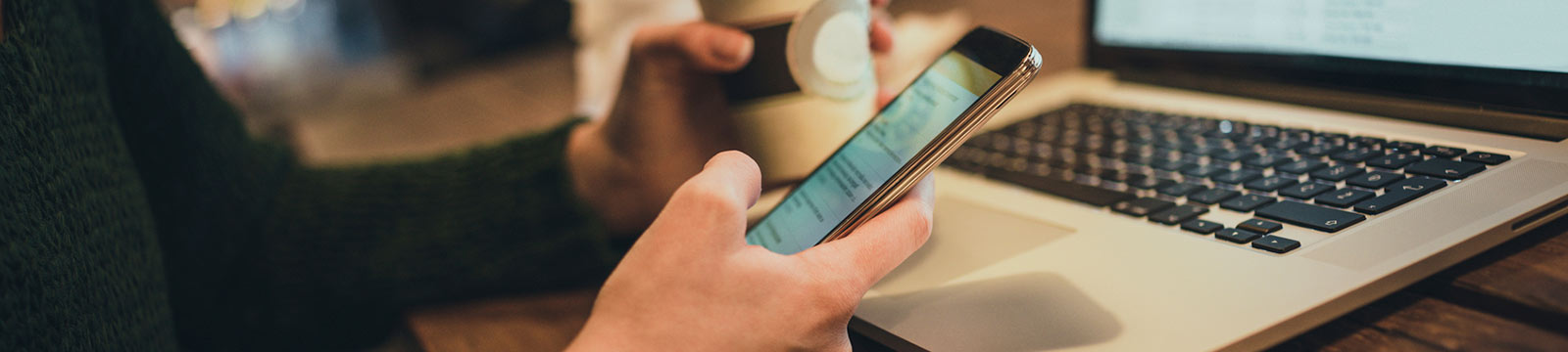 Woman holding phone and coffee in front of laptop.